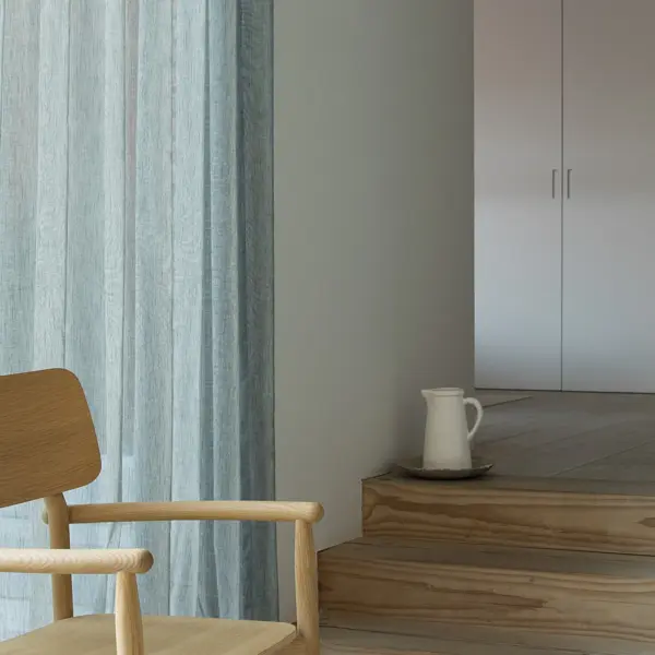 Forecast floor-to-ceiling sheer curtain fabric made from 100% Trevira CS polyester, featuring an inbetween transparent weave with subtle yarn thickening, shown in a minimalist modern interior with wooden steps, neutral walls, and soft daylight creating a calm, airy atmosphere, OEKO-TEX certified.