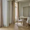 Hulst sheer curtain fabric in a soft neutral tone, made from a blended mix of acrylic, polyester, cotton, linen, and viscose, shown as inbetween transparent floor-length curtains in an elegant dining room with wooden flooring, minimalist furniture, and tall windows allowing warm natural light, OEKOTEX certified.