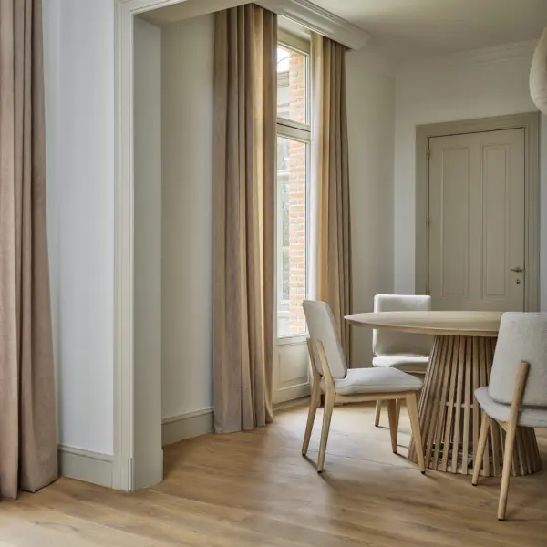 Hulst sheer curtain fabric in a soft neutral tone, made from a blended mix of acrylic, polyester, cotton, linen, and viscose, shown as inbetween transparent floor-length curtains in an elegant dining room with wooden flooring, minimalist furniture, and tall windows allowing warm natural light, OEKOTEX certified.