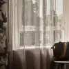 Lace sheer curtain fabric with a refined lino-style weave and subtle metallic sheen, made from 100% polyester, shown as inbetween transparent drapery in a sophisticated living room with large windows, soft natural light, and rich layered textures creating a luxurious interior atmosphere.