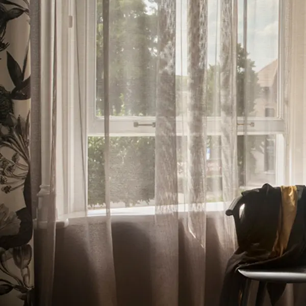 Lace sheer curtain fabric with a refined lino-style weave and subtle metallic sheen, made from 100% polyester, shown as inbetween transparent drapery in a sophisticated living room with large windows, soft natural light, and rich layered textures creating a luxurious interior atmosphere.