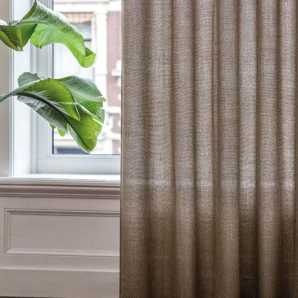 Mangaan sheer curtain fabric in a warm neutral tone, made from acrylic and polyester blend, shown as inbetween transparent floor-length curtains in a bright modern room with a large window, white wall paneling, wooden flooring, and indoor greenery filtering soft natural daylight, OEKOTEX certified.