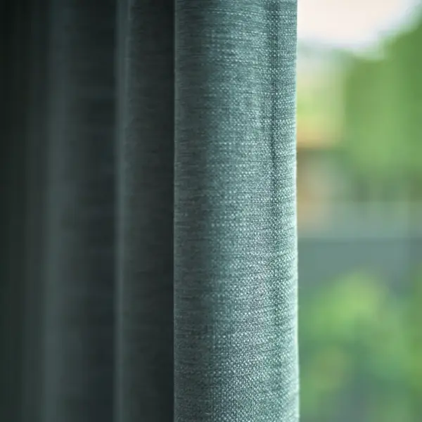 Nolin curtain fabric with an irregular woven texture and soft green-blue tone, made from recycled flame-retardant polyester, shown as floor-length drapery in a calm modern interior with large windows and greenery outside, creating a relaxed and natural atmosphere.