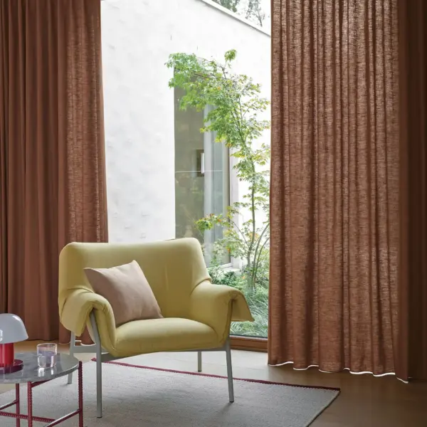 Ode-recycle curtain fabric with a closed weave texture, made from recycled and virgin Trevira CS polyester, shown as floor-to-ceiling drapery in a contemporary living room with large glass doors, indoor greenery, warm-toned curtains, and a modern lounge chair creating a cozy, design-led interior, OEKOTEX certified.