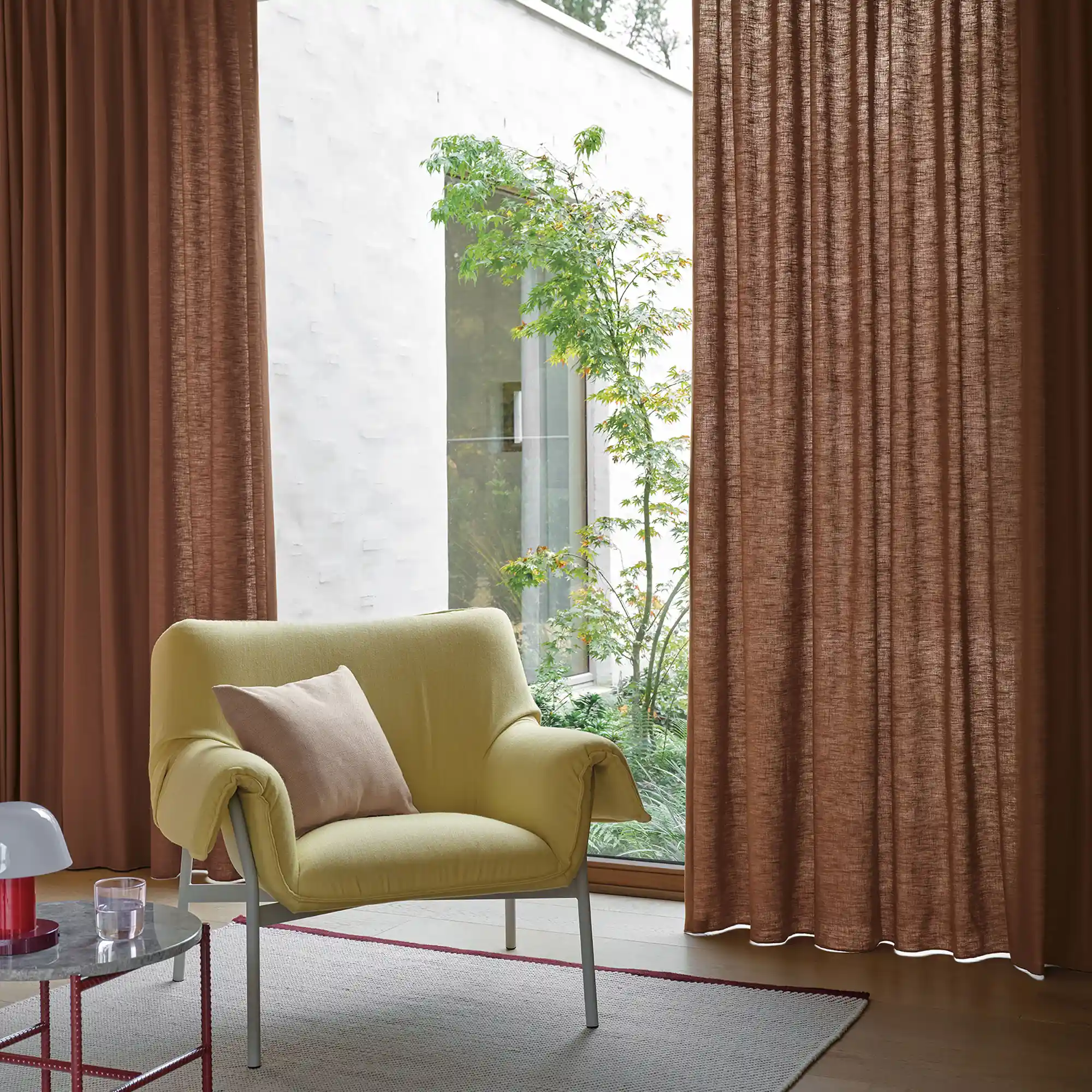 Ode-recycle curtain fabric with a closed weave texture, made from recycled and virgin Trevira CS polyester, shown as floor-to-ceiling drapery in a contemporary living room with large glass doors, indoor greenery, warm-toned curtains, and a modern lounge chair creating a cozy, design-led interior, OEKOTEX certified.