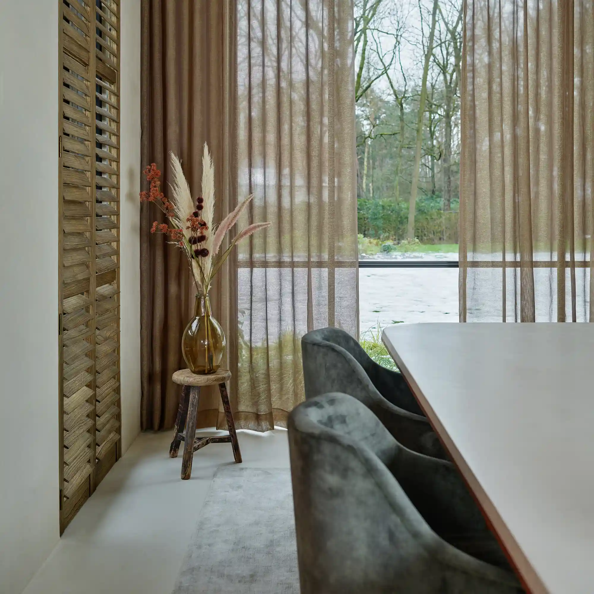 Oleander sheer curtain fabric with a refined closed structure, made from 100% flame-retardant polyester, shown as floor-to-ceiling drapery in a modern dining room with large windows, soft neutral furnishings, wooden accents, and natural daylight creating a calm, elegant atmosphere, OEKOTEX certified.