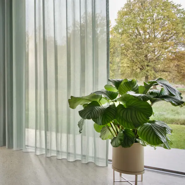 Onno sheer curtain fabric with inbetween transparent weave, made from a sustainable blend of cotton and recycled polyester, shown as floor-to-ceiling curtains in a bright modern living space with large glass doors, indoor plants, soft neutral flooring, and abundant natural light, OEKOTEX certified.