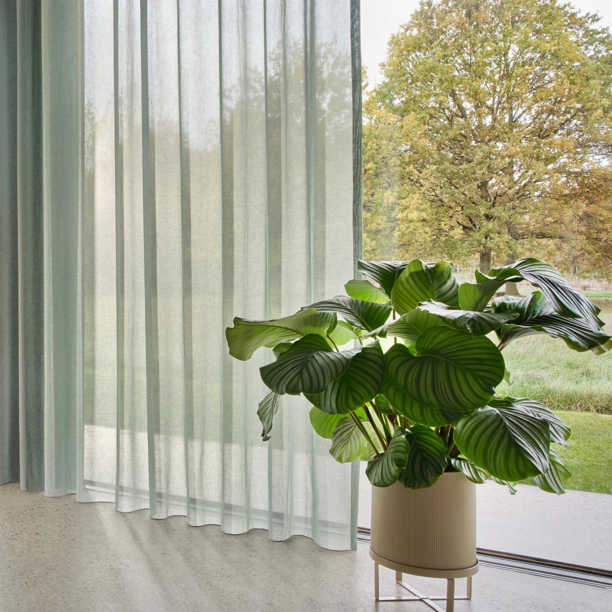 Onno sheer curtain fabric with inbetween transparent weave, made from a sustainable blend of cotton and recycled polyester, shown as floor-to-ceiling curtains in a bright modern living space with large glass doors, indoor plants, soft neutral flooring, and abundant natural light, OEKOTEX certified.