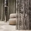 Opus sheer curtain fabric with inbetween transparent texture, made from a polyester and viscose blend, shown as layered floor-to-ceiling drapery in a contemporary living space with sculptural furniture, soft neutral upholstery, polished metal accents, and warm ambient lighting, OEKOTEX certified.
