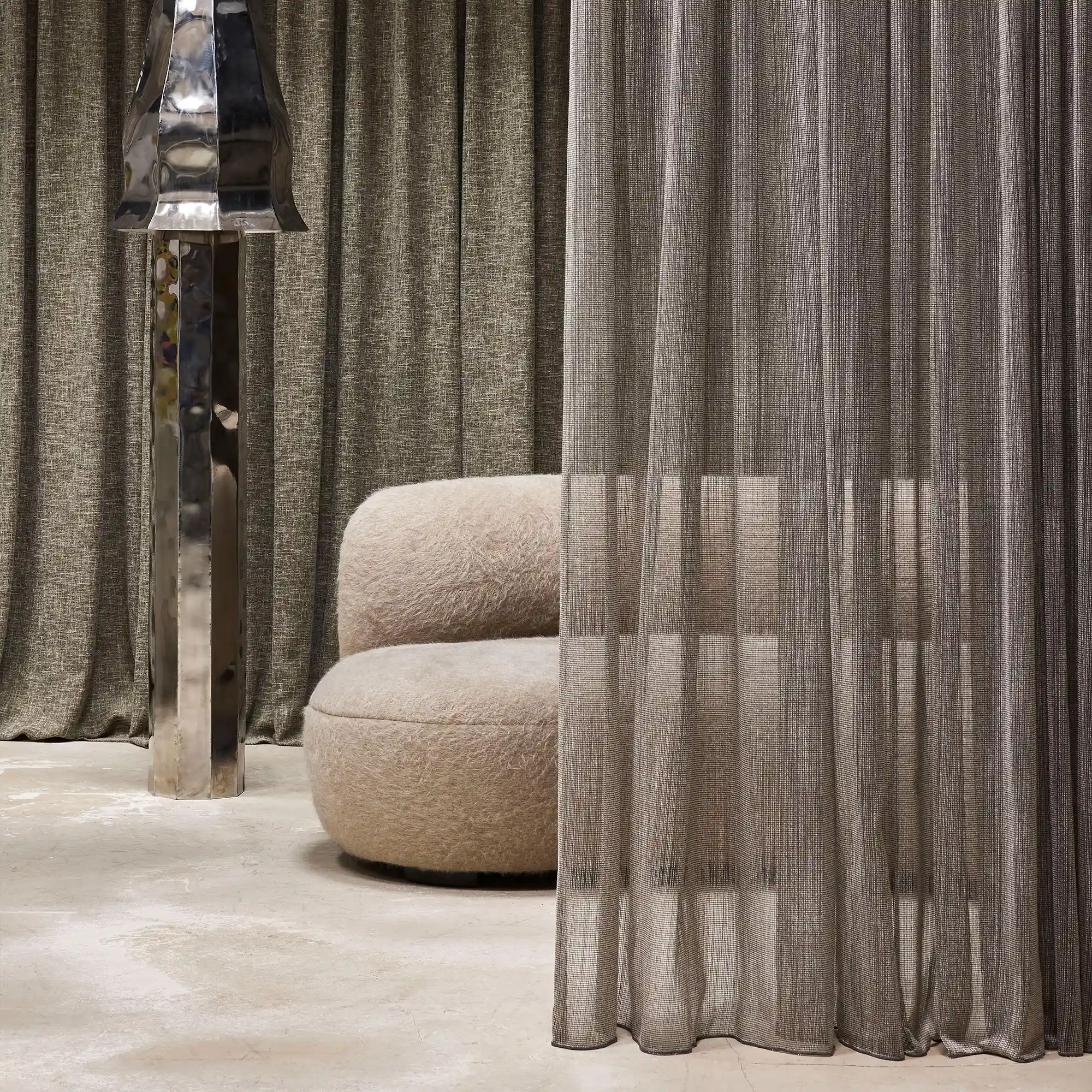 Opus sheer curtain fabric with inbetween transparent texture, made from a polyester and viscose blend, shown as layered floor-to-ceiling drapery in a contemporary living space with sculptural furniture, soft neutral upholstery, polished metal accents, and warm ambient lighting, OEKOTEX certified.